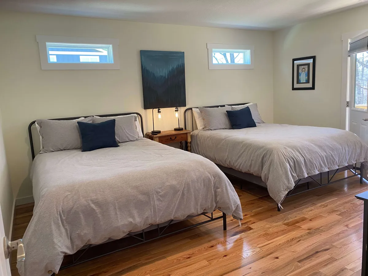 A tranquil bedroom with two beds dressed in light gray bedding, complemented by wall artwork and a wooden bedside table with lamps. The room feels spacious yet cozy, making it suitable for families or friends sharing the space.