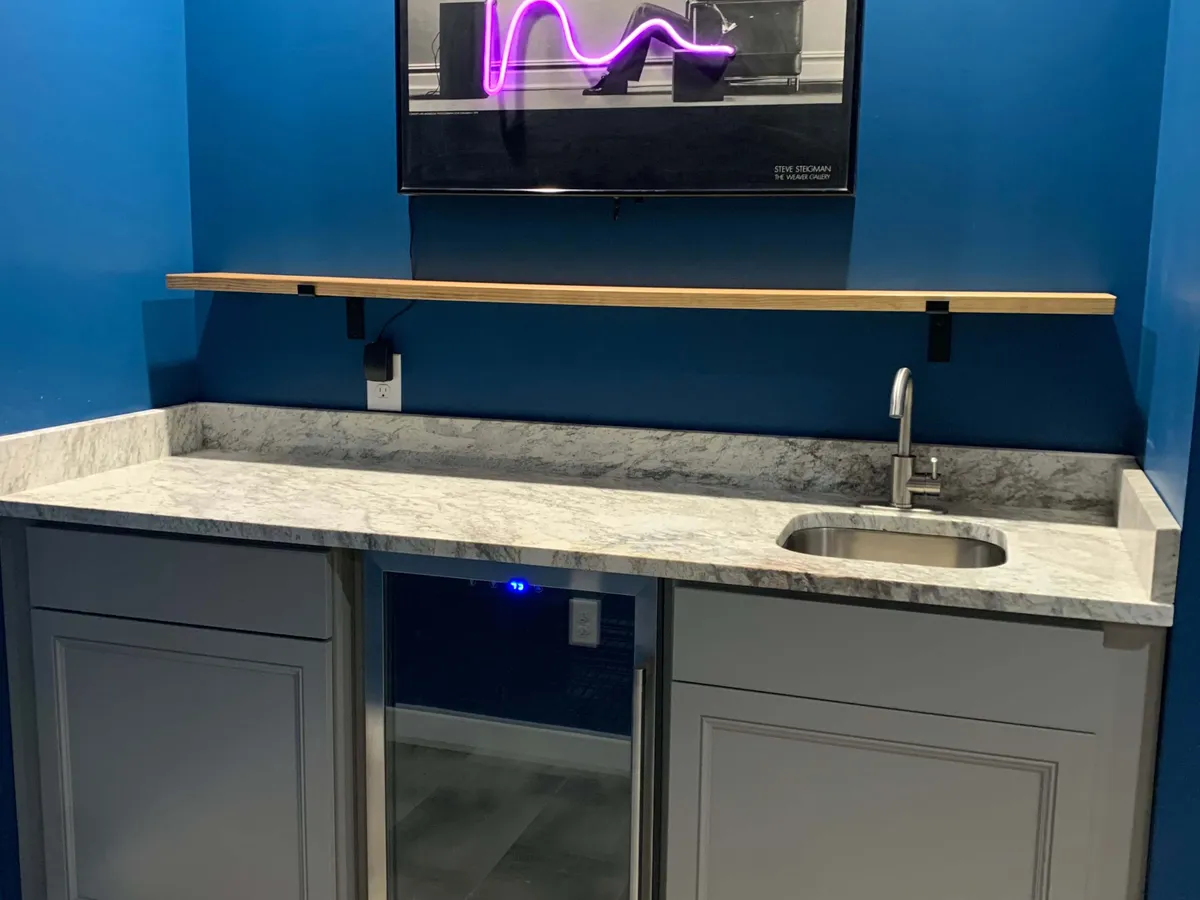 A vibrant kitchen scene with striking blue walls, a long marble countertop, a wooden shelf stocked for entertaining, a sink, and a neon art piece. The space is lively and inviting, designed for both culinary creativity and social gatherings.