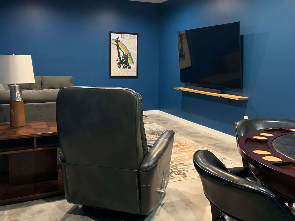 A comfortable lounge area featuring a gray sofa, reclining chair, large black television, a wooden coffee table, and colorful wall art. The space is well-lit and designed for group activities like watching movies, playing games, or relaxing with friends.