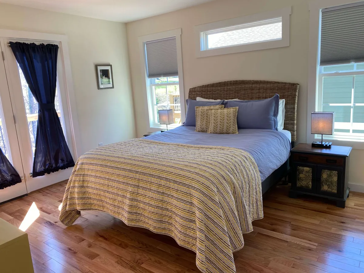 A serene bedroom scene featuring a bed with patterned bedding, decorative pillows, a nightstand with a lamp, and windows adorned with curtains. The natural light filters softly into the room, creating a peaceful environment ideal for relaxation or enjoying a quiet morning coffee.