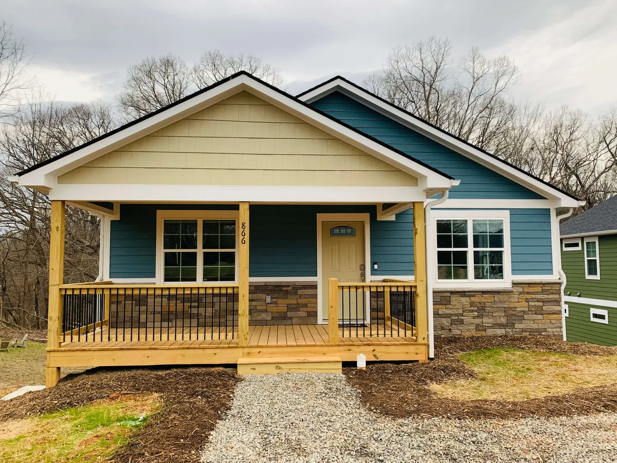 A charming blue bungalow with inviting white trim, a wide front porch perfect for gathering, and a sturdy stone foundation. The porch overlooks a lush yard, capturing the essence of relaxed Southern living in Asheville.